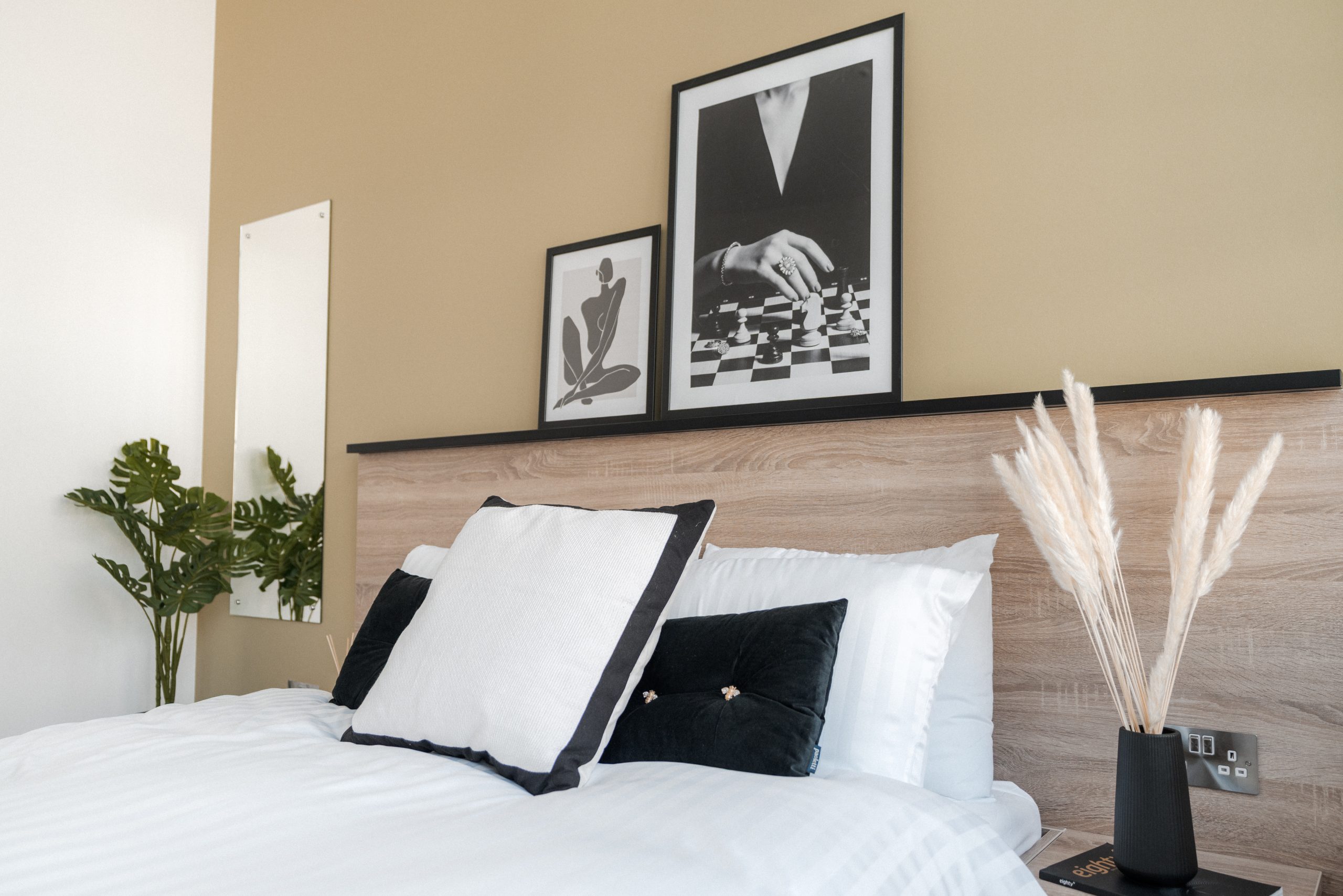 Bed inside YourTRIBE's Deptford student accommodation, with white and black bedding, pampas grass in a black vase on a bedside table, wall mirror, plants, and images in frames.