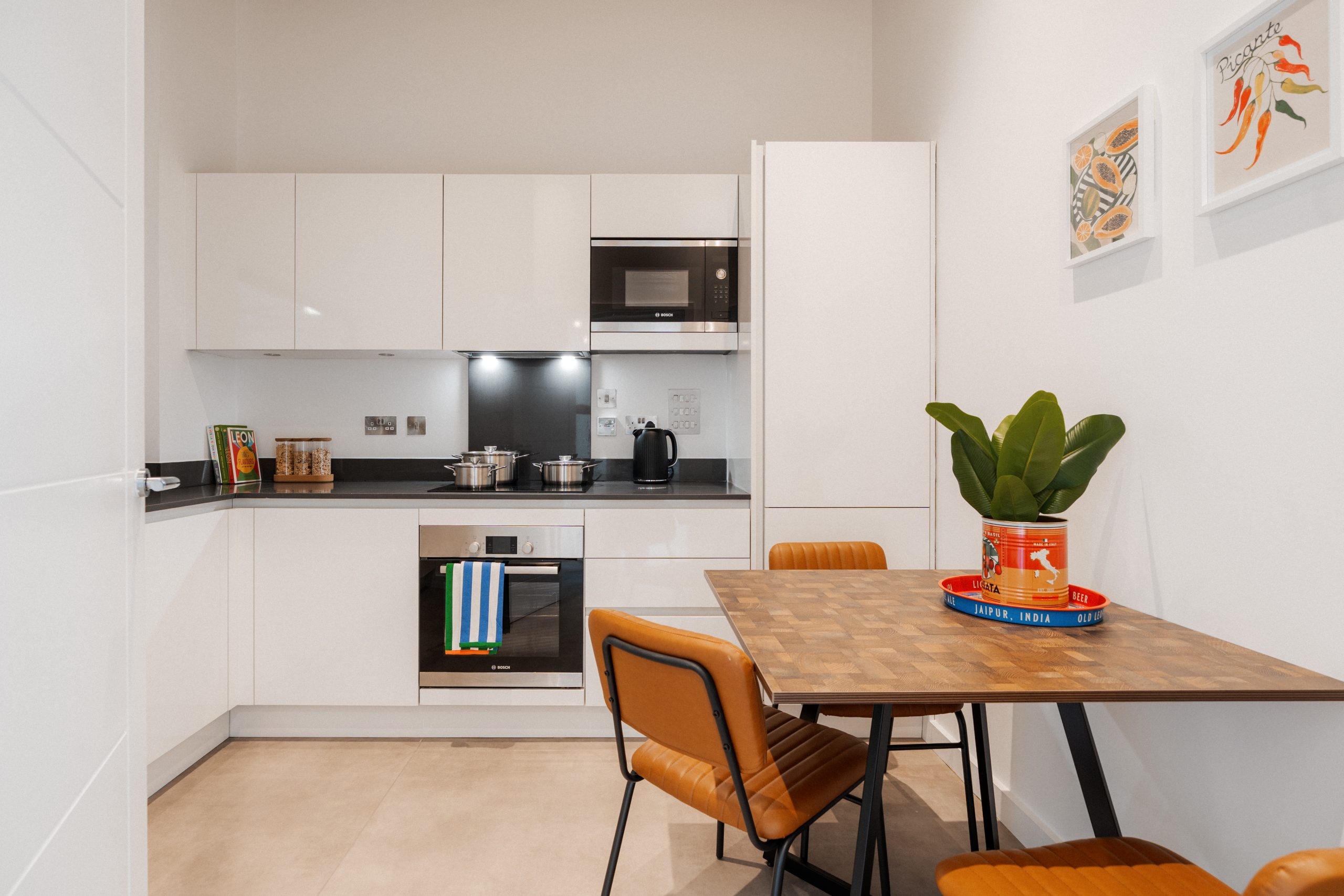 Kitchen dining area inside YourTRIBE's Deptford student accommodation. A white kitchen with a wooden dining table and orange chairs.